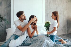 A husband surprising his wife for Mother's Day- their daughter is holding flowers to give her.